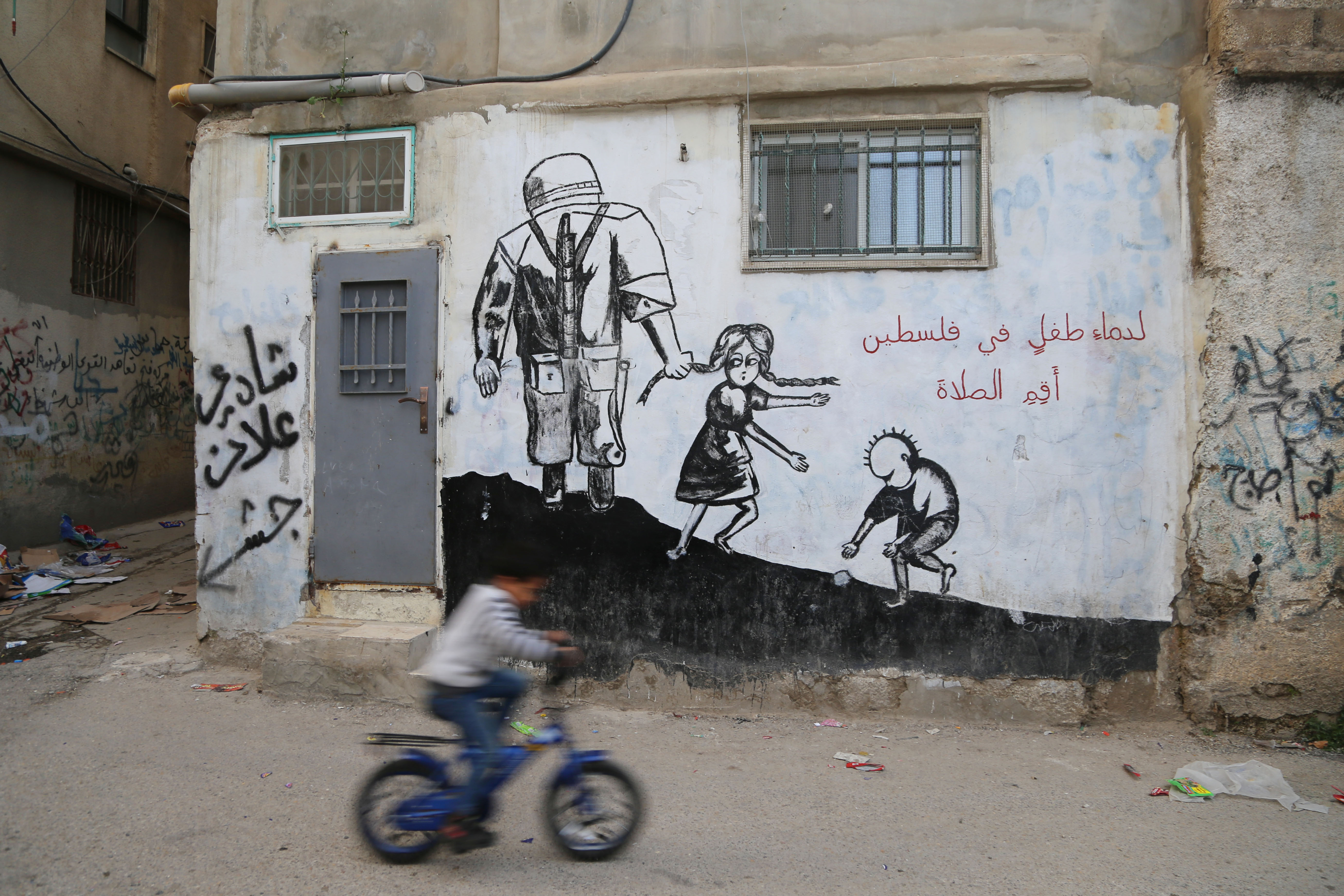 A child bicycles by a mural in Aida refugee camp. Aida is one of the 58 recognized refugee camps established after 1948 to accommodate Palestinians who were expelled in the 1948 Palestinian exodus. Keys are a popular motif of the camp, because many of the camp’s refugees still have their keys to their 1948 homes. Aida means “one who will return.”